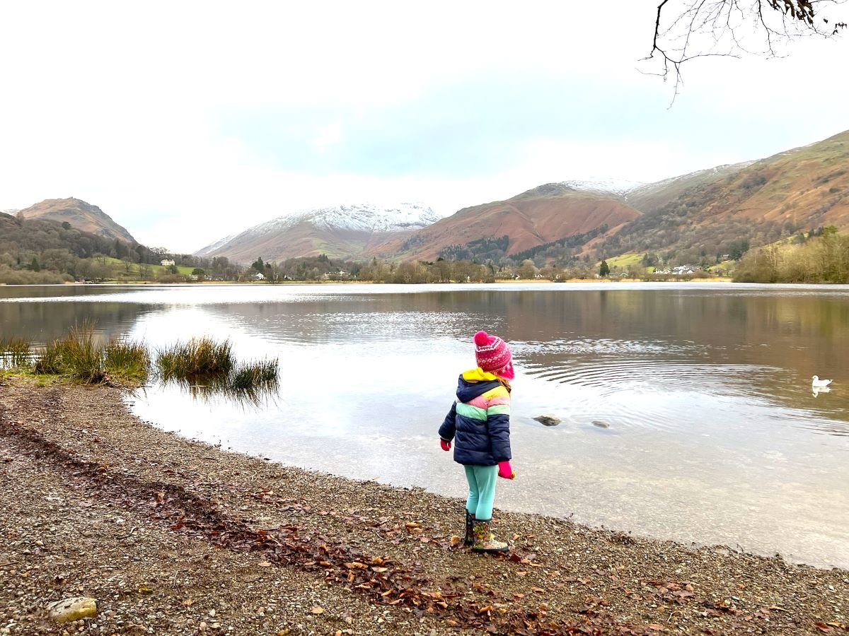 A small child in a stripy puffy coat and wellies looks out at Elterwater.