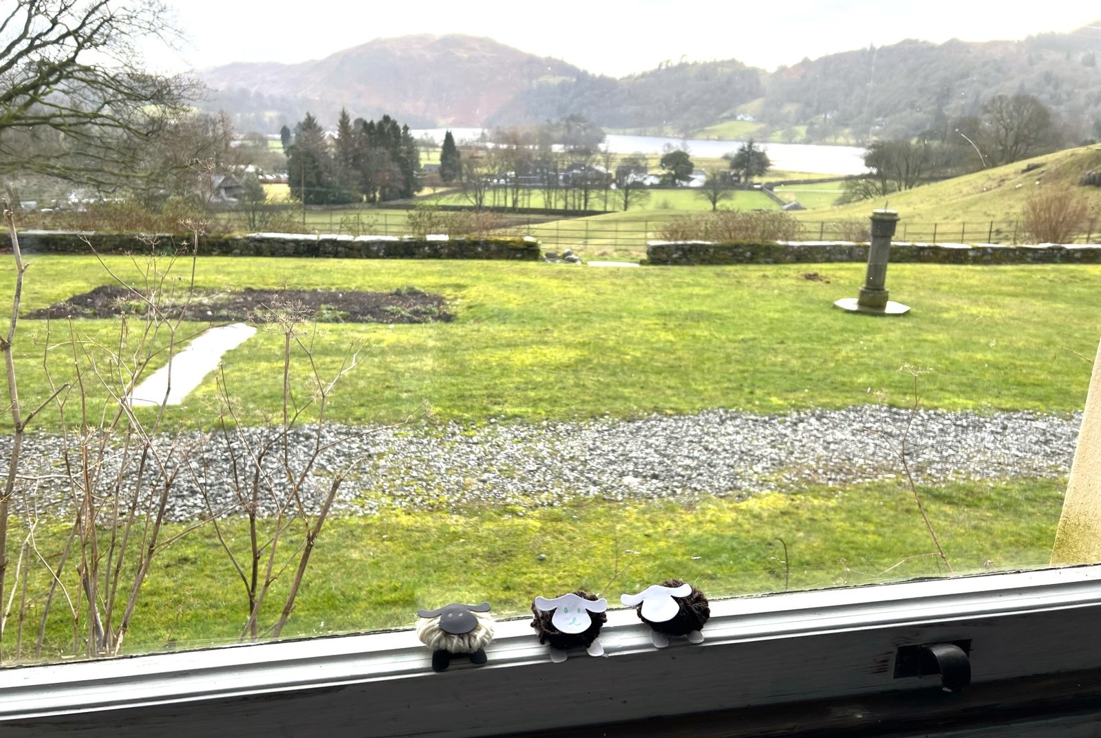 Three crafted woolly sheep sit on the window of Allan Bank. The view is of Grasmere lake and surrounding fells.