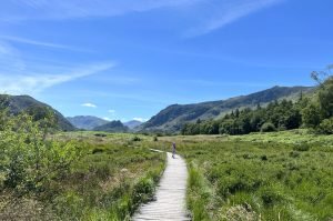 boardwalk round derwentwater, with mountains in the background. The sky is blue and a child skips along the boardwalk.
