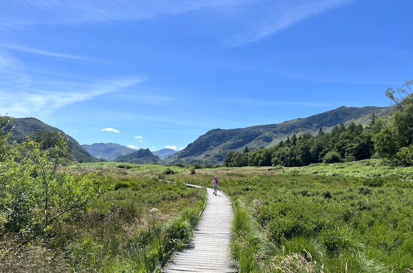 boardwalk round derwentwater, with mountains in the background. The sky is blue and a child skips along the boardwalk.