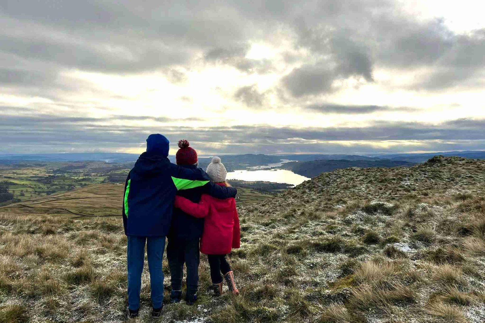 Three children are facing away from the camera, looking out over Lake Windermere. They are standing on Wansfell Pike