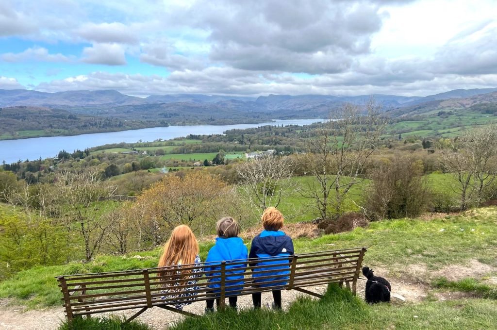 Three children sit on a bench with their backs to the camera. They look out over Lake Windermere from the top of Orrest Head.