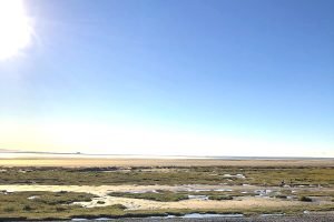 Bardsea Beach which is flat with grassy areas. The sky is clear and blue and the sun is in the sky.