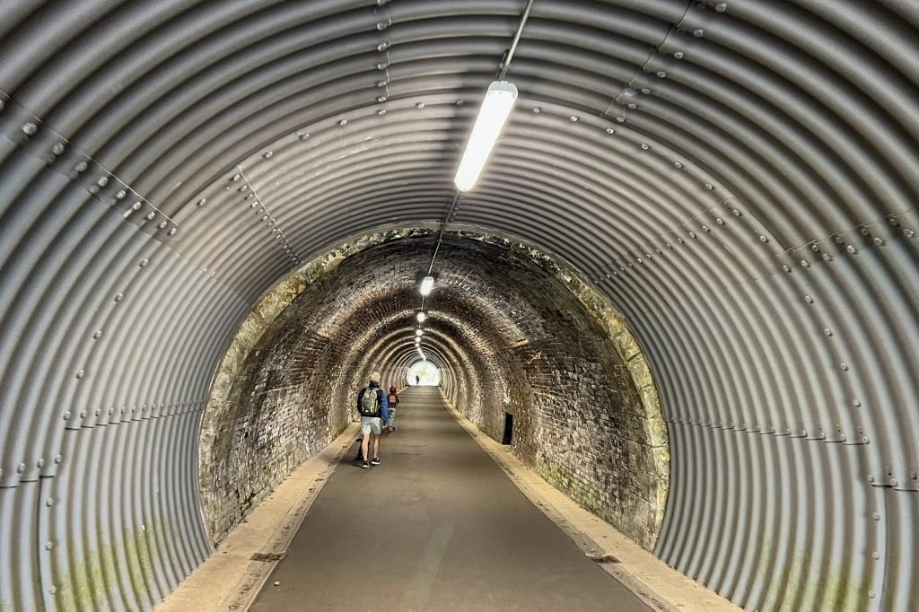 A man walks through the Keswick to Threlkeld Tunnel. In the distance two children are running and scooting out of the tunnel.