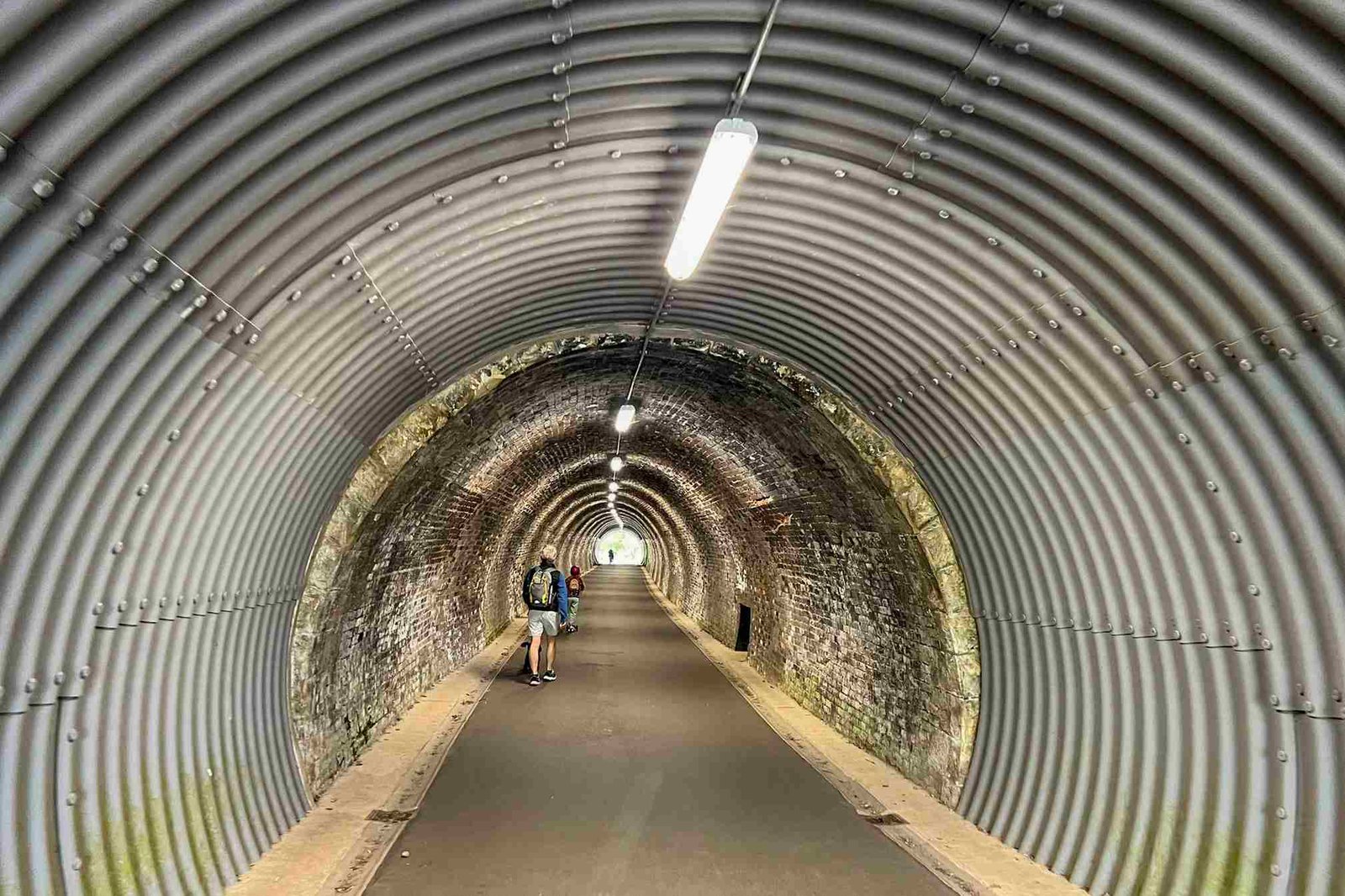 A man walks through the Keswick to Threlkeld Tunnel. In the distance two children are running and scooting out of the tunnel.