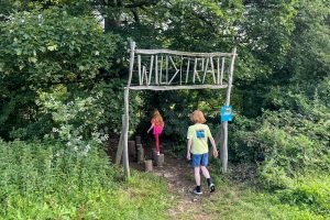 Two children walk under a sign to enter the Wild Play Trail at Sizergh. The sign says Wild Trail and is all made of sticks.