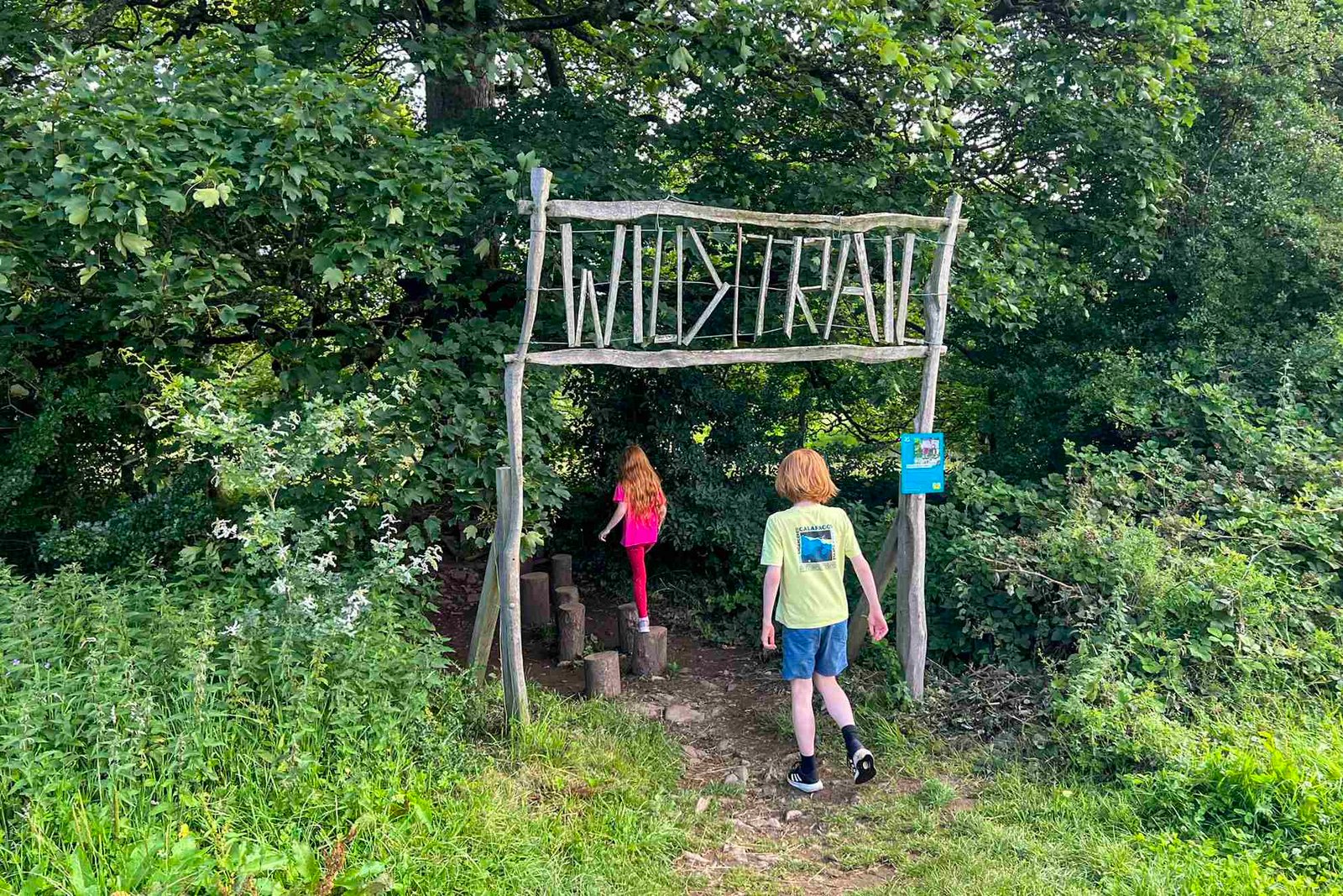 Two children walk under a sign to enter the Wild Play Trail at Sizergh. The sign says Wild Trail and is all made of sticks.